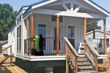 Cozy tiny house with white siding and wooden accents, featuring a small porch with potted plants. The surrounding area is bright and sunny, evoking a welcoming, serene vibe.