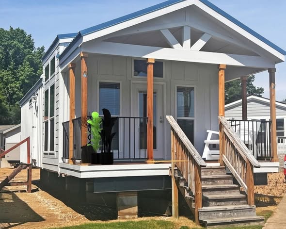 Cozy tiny house with white siding and wooden accents, featuring a small porch with potted plants. The surrounding area is bright and sunny, evoking a welcoming, serene vibe.