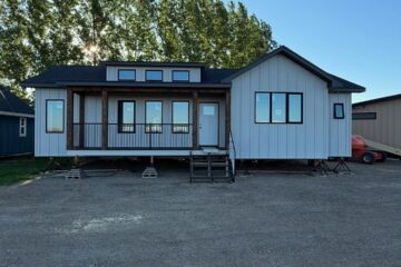 A modern, white modular home with black accents, set on a gravel lot against a backdrop of tall trees under a clear blue sky. Calm and inviting atmosphere.