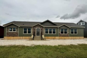 Single-story green house with a stone accent facade, surrounded by gravel. Overcast sky and grassy foreground create a calm, suburban feel.