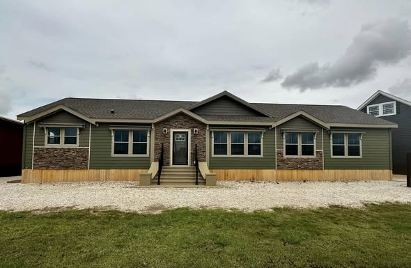Single-story green house with a stone accent facade, surrounded by gravel. Overcast sky and grassy foreground create a calm, suburban feel.