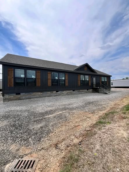 A modern, dark gray house with large windows and wooden shutters sits on a gravel path. The sky above is partly cloudy, adding a serene tone.
