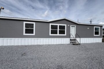 A gray mobile home with white trim sits on a gravel lot under a cloudy sky. It features three windows, a white door, and a metal staircase.