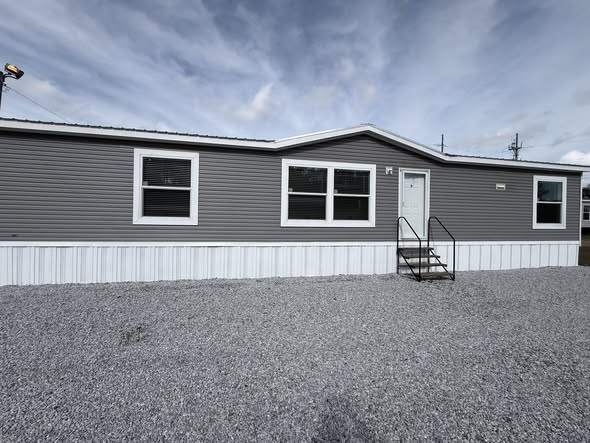 A gray mobile home with white trim sits on a gravel lot under a cloudy sky. It features three windows, a white door, and a metal staircase.