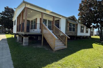Single-story white house on raised foundation, with wooden porch and steps. Bright sun and clear sky, casting shadows on the green lawn.