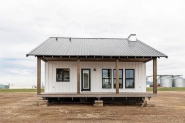 Small white modern farmhouse with a metal roof, wooden posts, and front porch. It stands on a rural, open field with silos in the background.