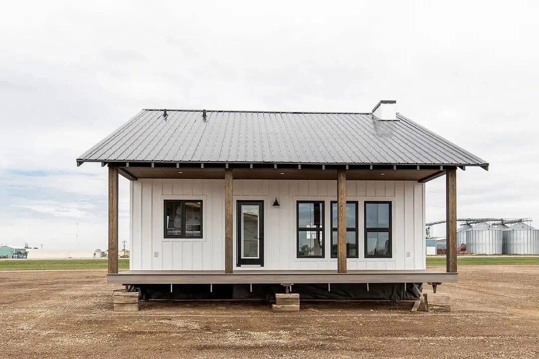 Small white modern farmhouse with a metal roof, wooden posts, and front porch. It stands on a rural, open field with silos in the background.