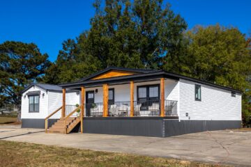 A modern white mobile home with a wooden porch and steps sits under a clear blue sky. Large green trees in the background add a serene feel.