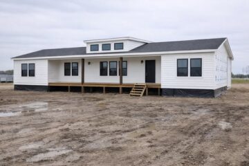 A white, single-story prefab house with a dark roof on a muddy, undeveloped lot. It features a porch with wooden steps and large windows, under a cloudy sky.