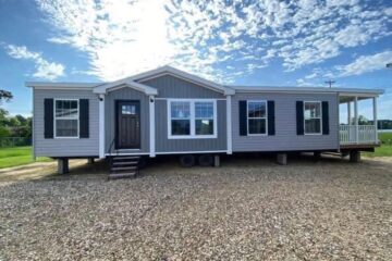 A modern, gray modular home with white trim and black shutters sits elevated on a gravel driveway under a bright blue sky with scattered clouds.