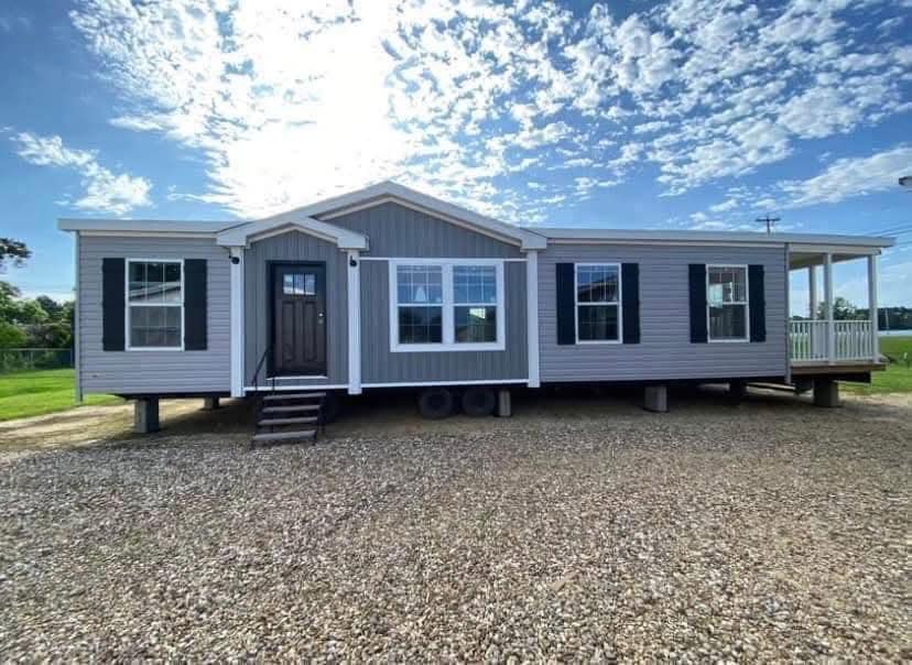 A modern, gray modular home with white trim and black shutters sits elevated on a gravel driveway under a bright blue sky with scattered clouds.