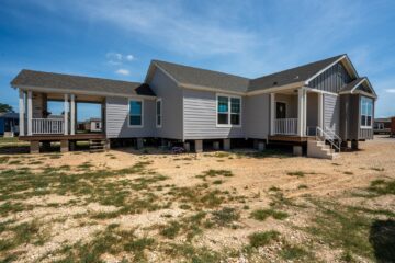 A gray manufactured home with multiple sections sits elevated on a dry, grassy lot under a blue sky. It features a small porch and white stairs.