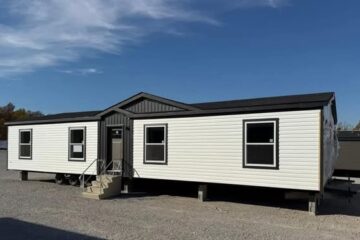 A white double-wide mobile home with a dark roof sits on a gravel lot under a clear blue sky. Three front steps lead to the main entrance.