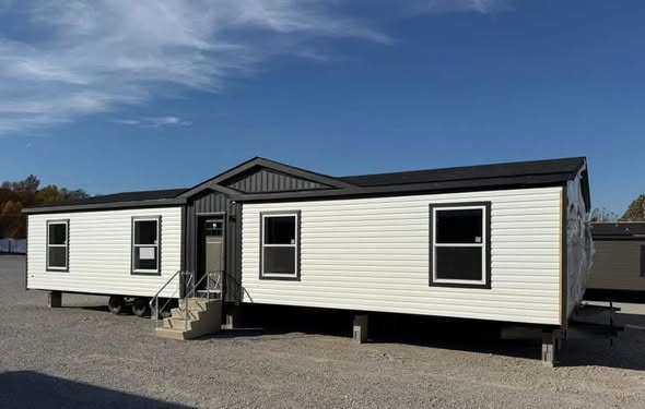 A white double-wide mobile home with a dark roof sits on a gravel lot under a clear blue sky. Three front steps lead to the main entrance.