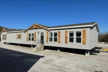 A modern, gray manufactured home with wooden shutters and trim sits elevated on a gravel lot. The bright blue sky accentuates its clean lines and inviting exterior.