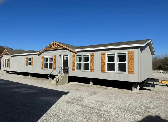 A modern, gray manufactured home with wooden shutters and trim sits elevated on a gravel lot. The bright blue sky accentuates its clean lines and inviting exterior.