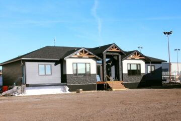 Single-story house with dark siding and gable roofs, surrounded by a dirt yard on a clear day. A small staircase leads to the main entrance.