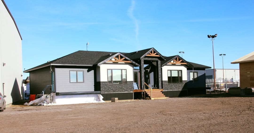 Single-story house with dark siding and gable roofs, surrounded by a dirt yard on a clear day. A small staircase leads to the main entrance.