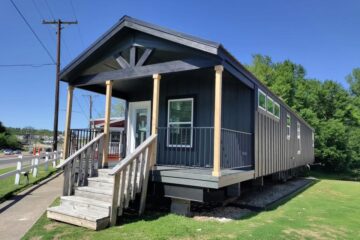 A dark-colored tiny house on a wooden foundation with a small porch and stairs. It's set beside a street, surrounded by green grass and trees, under a clear blue sky.