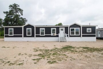 A black and white mobile home with five windows and a central door, surrounded by a gravel yard and trees against a cloudy sky.