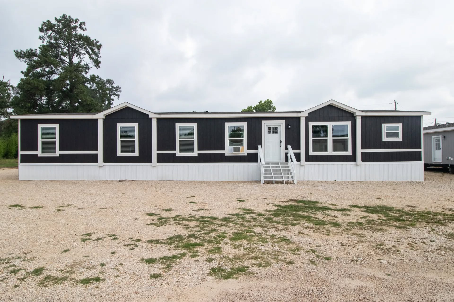 A black and white mobile home with five windows and a central door, surrounded by a gravel yard and trees against a cloudy sky.