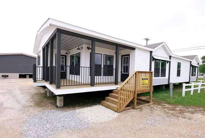 Manufactured home with a spacious, elevated front porch, black railings, and white siding. Wooden steps lead up, surrounded by gravel and grass, under a cloudy sky.