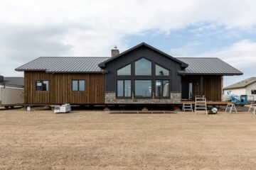 Modern house under construction with wooden siding, large windows, and a metal roof. The setting is a dirt lot with a cloudy sky, conveying progress.