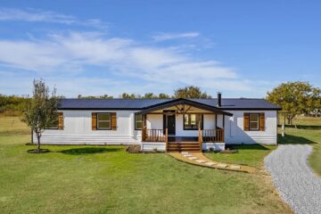A white ranch-style house with wooden shutters and a front porch sits on a vast green lawn. A gravel path leads to the entrance. The sky is clear and blue.