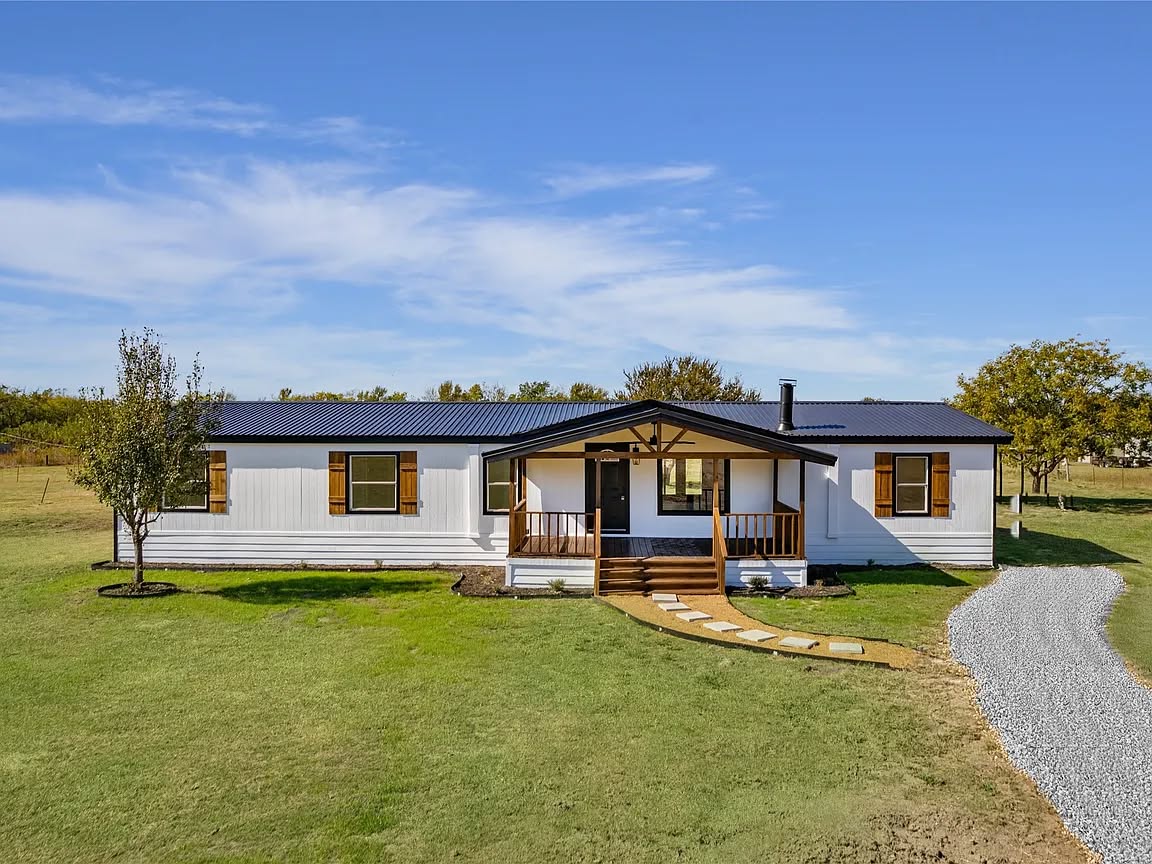 A white ranch-style house with wooden shutters and a front porch sits on a vast green lawn. A gravel path leads to the entrance. The sky is clear and blue.