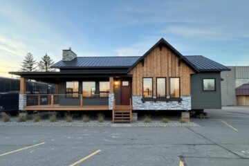 A modern single-story house with wooden and stone facade, large windows, and a gable roof. It has a porch with railing and is set against a clear sky.