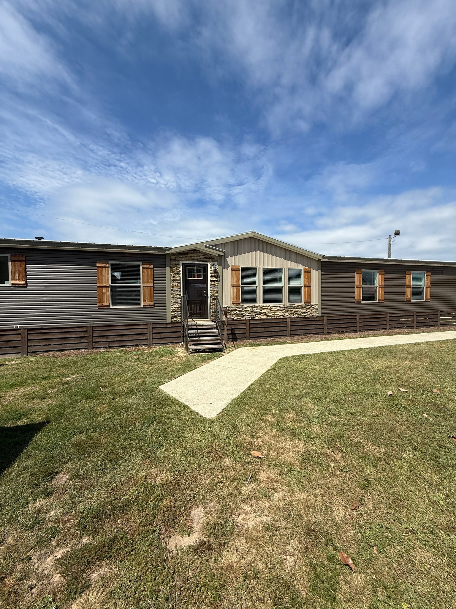A single-story manufactured home with stone accents and wooden shutters sits on a grassy lawn under a blue sky with wispy clouds. A concrete path leads to the entrance.