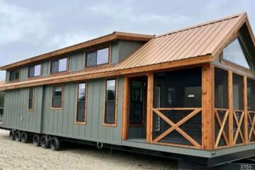 A modern tiny house on wheels with green siding and a wood-trimmed roof. Large windows and a front porch create a cozy, inviting atmosphere.