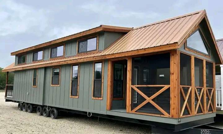 A modern tiny house on wheels with green siding and a wood-trimmed roof. Large windows and a front porch create a cozy, inviting atmosphere.
