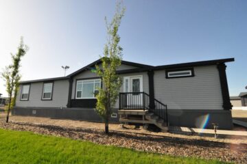 Single-story modular home with gray siding and black trim, set against a clear blue sky. Surrounded by young trees and a neatly landscaped yard.