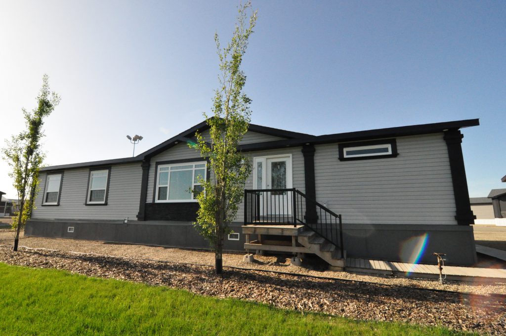 Single-story modular home with gray siding and black trim, set against a clear blue sky. Surrounded by young trees and a neatly landscaped yard.