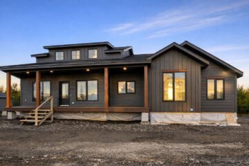 Single-story modern home with dark wood siding, large windows, and a covered porch. Set against a clear evening sky, creating a serene atmosphere.