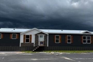 A long, single-story dark gray building with a white roof and wooden shutters sits under a dramatic, cloudy sky, suggesting an approaching storm.
