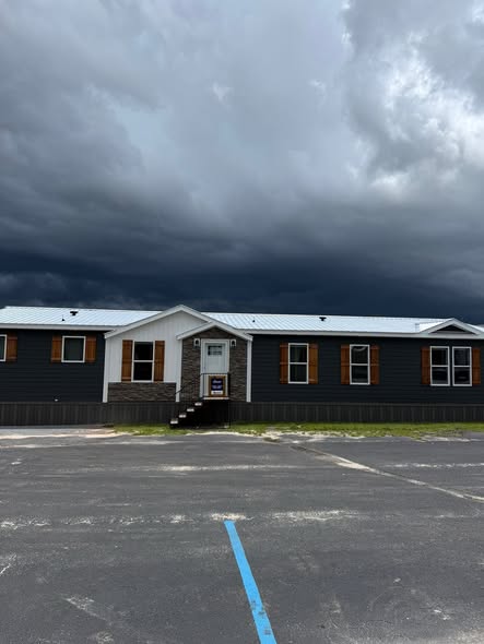 A long, single-story dark gray building with a white roof and wooden shutters sits under a dramatic, cloudy sky, suggesting an approaching storm.