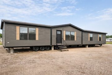 A dark gray mobile home with three visible sections, resting on a gravel lot under a clear blue sky. It features brown shutters and a central door with stairs.
