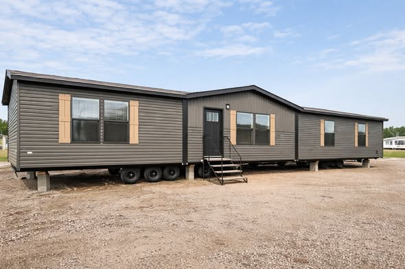 A dark gray mobile home with three visible sections, resting on a gravel lot under a clear blue sky. It features brown shutters and a central door with stairs.