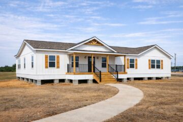 A white modular home with a gray roof and wooden shutters sits elevated on a dry lawn. A curved concrete path leads to its porch, giving a welcoming feel.