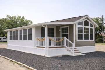 A small, single-story mobile home with a gabled roof, featuring large windows and a porch adorned with a garland. The house is set on a gravel lot.