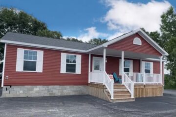 A charming single-story house with red siding and white trim sits under a bright blue sky. It features a gabled entrance, white railing, and stone foundation.