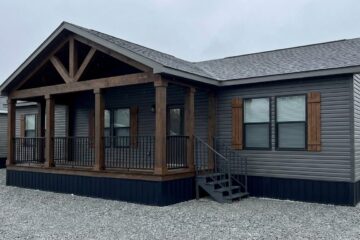 A modern, gray modular home with a wooden porch and gabled roof. The inviting exterior features large windows and wooden shutters, set on a gravel lot.
