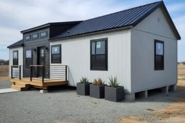 Small modern house with gray paneling and black roof under a clear blue sky. Front porch has railings and three large potted plants. Sparse surroundings.