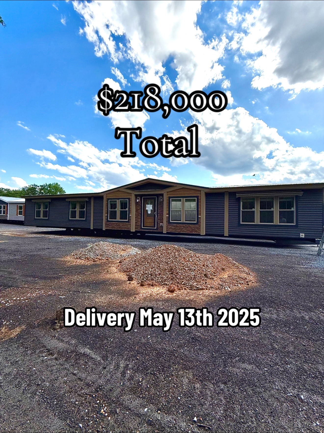 Single-story modular home under blue sky with clouds, priced at $218,000, and scheduled for delivery on May 13, 2025. Pile of dirt in foreground.