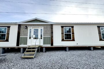 Modern mobile home with a white exterior and wooden accents, featuring a central entrance with steps and two large windows, set on gravel.