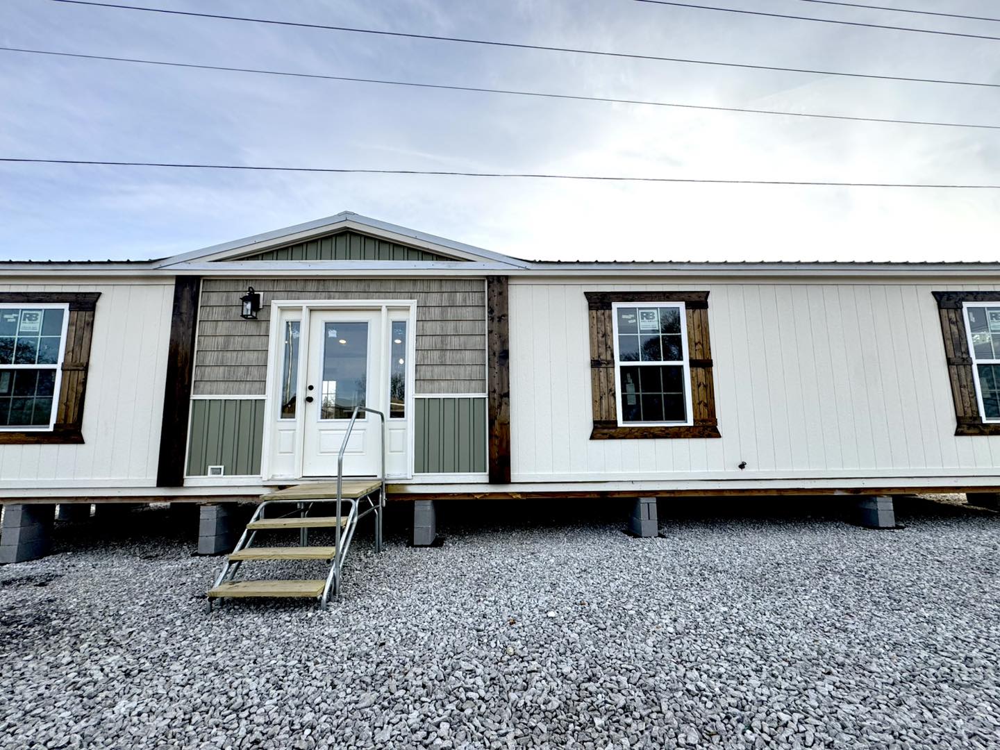 Modern mobile home with a white exterior and wooden accents, featuring a central entrance with steps and two large windows, set on gravel.