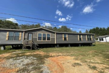 A long, dark gray mobile home with wooden shutters sits on a grassy lot under a blue sky with fluffy clouds, evoking a sense of simplicity and openness.