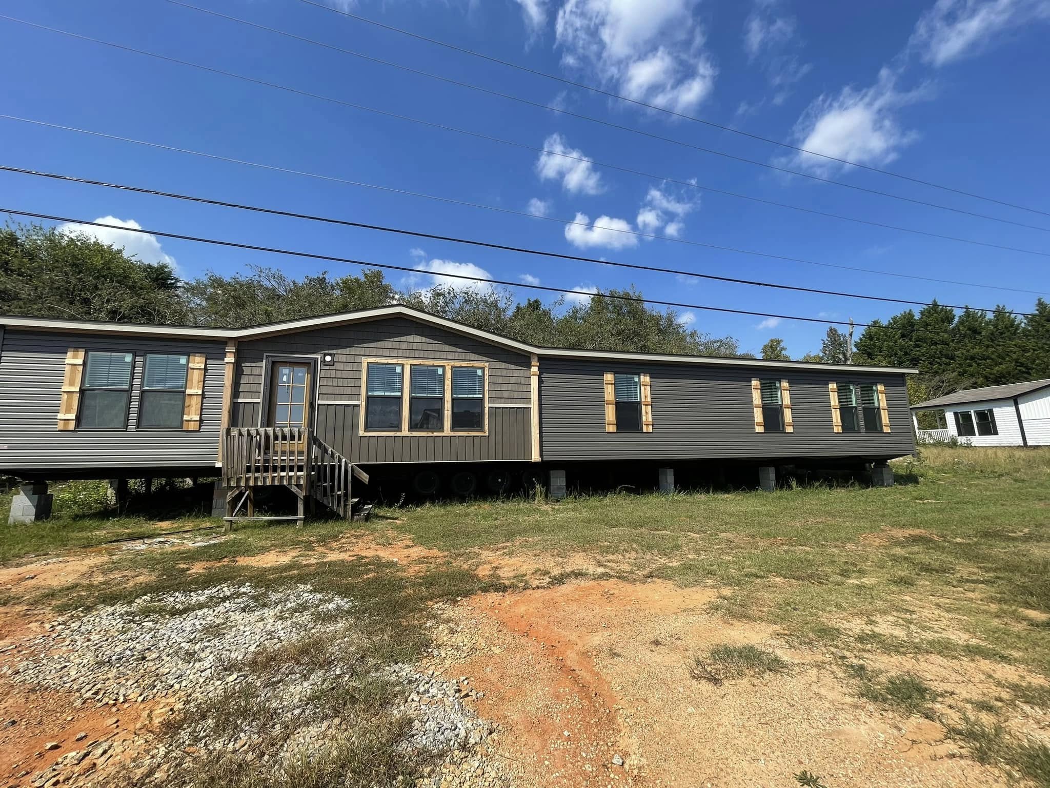 A long, dark gray mobile home with wooden shutters sits on a grassy lot under a blue sky with fluffy clouds, evoking a sense of simplicity and openness.